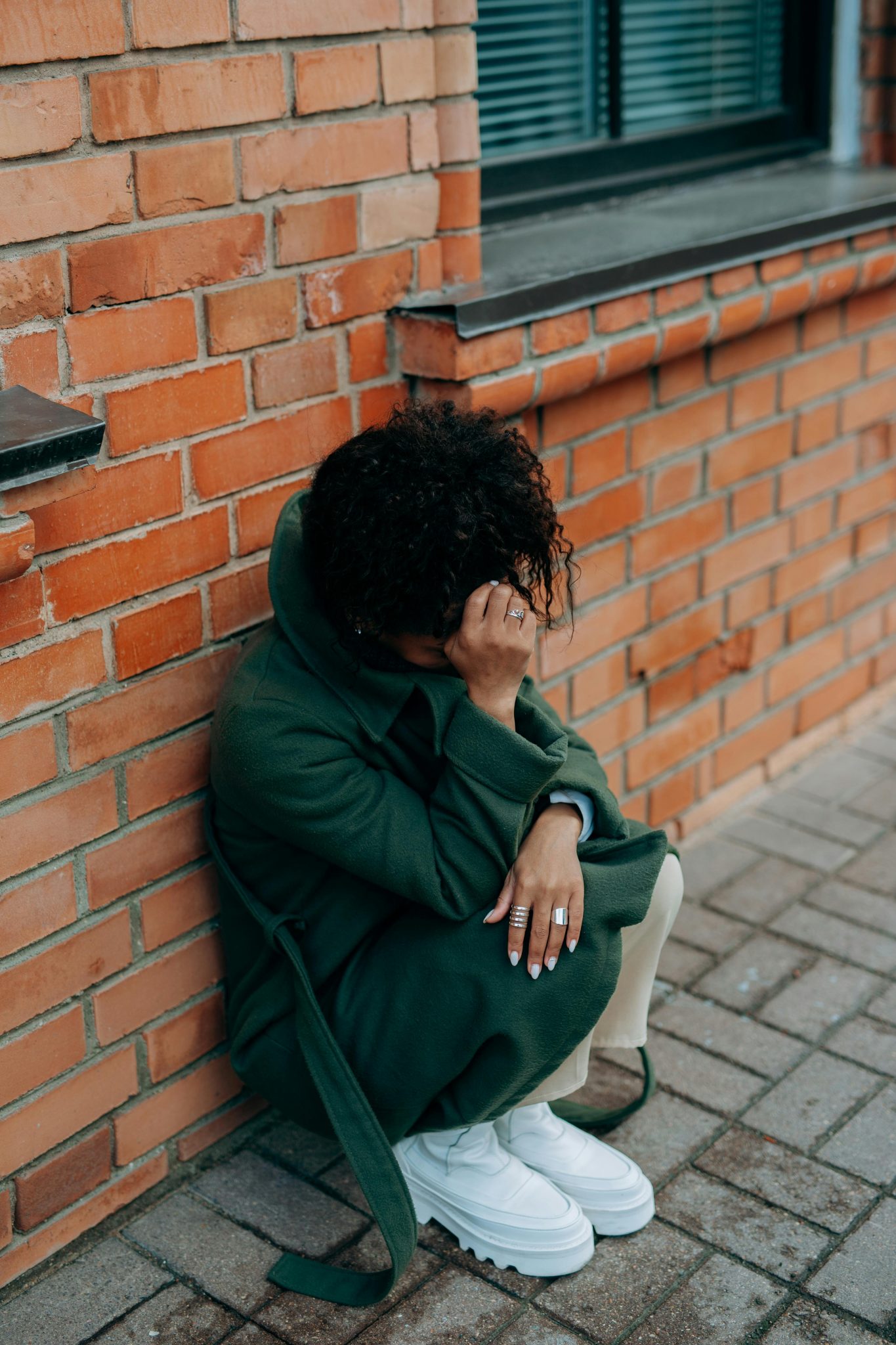 A woman in a green coat sits against a brick wall, deep in thought.